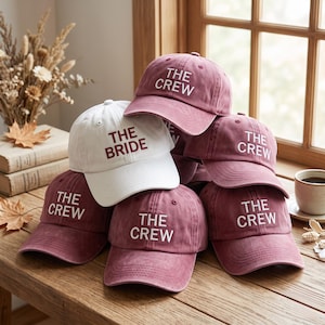 May include: A collection of baseball caps stacked together. One white cap features the text THE BRIDE in dark red embroidery. Five dusty rose-coloured caps feature the text THE CREW in white embroidery. Each cap has a curved brim and eyelets.