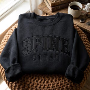 May include: A black long-sleeve crewneck sweatshirt featuring the text SPINE SQUAD embroidered in a matching black thread across the chest. The word SPINE is arched in a bold collegiate font, while the word SQUAD is in a smaller, straight font.