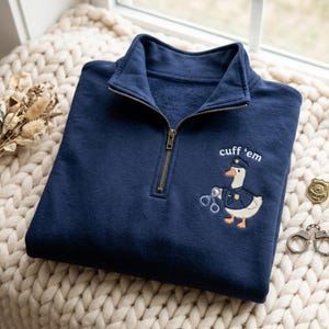 May include: A navy blue quarter-zip sweatshirt featuring an embroidered graphic of a white goose dressed as a police officer, holding a pair of handcuffs. Above the goose, the text reads "cuff 'em" in white, lowercase, sans-serif lettering.
