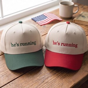 Può includere: Due cappellini da baseball beige a cinque pannelli con un bottone sulla parte superiore. Un cappellino presenta un ricamo verde scuro con la scritta "he's running" e visiera abbinata. L'altro ha un ricamo rosso "he's running" e visiera rossa.