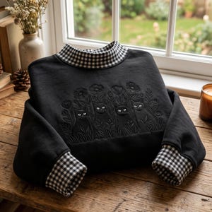 Puede incluir: Una sudadera negra de manga larga con cuello alto y puños en un patrón de cuadros vichy blanco y negro. La parte delantera de la prenda muestra un diseño en relieve de cuatro gatos negros entre diversas flores y follaje.