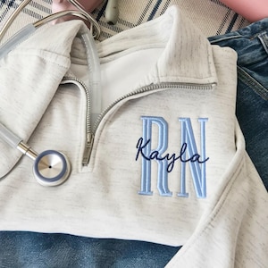 Pode incluir: Uma camisola cinza claro com fecho de correr de um quarto com as letras bordadas "RN" e o nome "Kayla" em azul claro. Um estetoscópio repousa sobre a camisola. A camisola tem uma gola branca e um fecho de correr prateado.