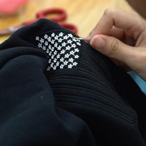 May include: A black fabric garment featuring a rectangular patch with a repeating white floral pattern. The patch is being worked on with a small metal tool, showing detailed stitching and a textured surface on the black material surrounding it.