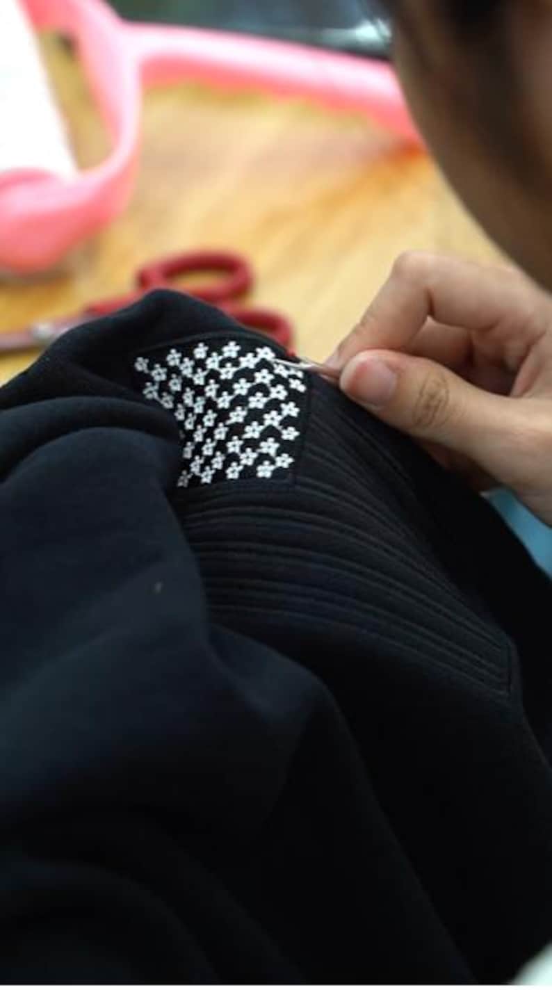 May include: A black fabric garment featuring a rectangular patch with a repeating white floral pattern. The patch is being worked on with a small metal tool, showing detailed stitching and a textured surface on the black material surrounding it.