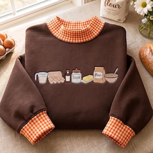 May include: A dark brown long-sleeve sweatshirt featuring an orange and white gingham collar and cuffs. The center of the chest displays an embroidered graphic of baking ingredients including a measuring cup, eggs, vanilla, sugar, butter, flour, and a bowl.
