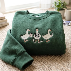 Op de afbeelding: Een donkergroene sweater met ronde hals en een geborduurde afbeelding van drie witte ganzen. De middelste gans houdt een echofoto vast, de linker gans een notitieblok en de rechter gans een injectiespuit. Medische flesjes en een test liggen eronder.