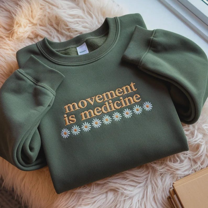 Pu&ograve; includere: Felpa verde oliva con la scritta ricamata "movement is medicine" in filo dorato, con una fila di margherite bianche e gialle. La felpa ha maniche lunghe e girocollo.