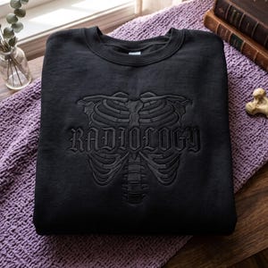 May include: A black crewneck sweatshirt featuring a tonal black embroidered design of a human rib cage and spine. The word RADIOLOGY is embroidered in a gothic-style font across the center of the rib cage, creating a subtle, monochromatic look.