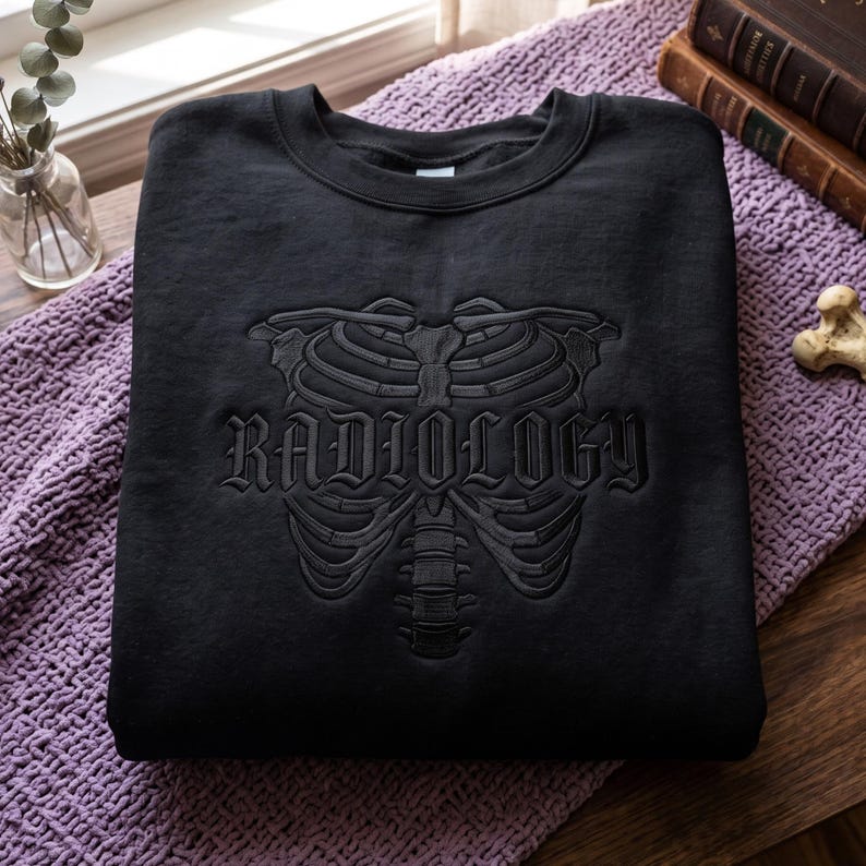 May include: A black crewneck sweatshirt featuring a tonal black embroidered design of a human rib cage and spine. The word RADIOLOGY is embroidered in a gothic-style font across the center of the rib cage, creating a subtle, monochromatic look.