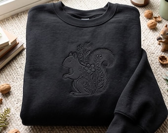 May include: A black crewneck sweatshirt featuring a tonal embroidered design on the front. The embroidery depicts a squirrel surrounded by intricate floral and botanical elements, all rendered in the same black thread as the fabric.