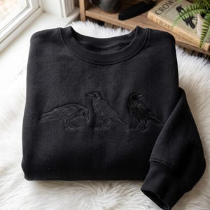 Könnte beinhalten: Ein schwarzes Sweatshirt mit Rundhalsausschnitt und einer tonalen Stickerei von drei Krähen auf der Brust. Das Kleidungsstück hat einen gerippten Kragen und gerippte Bündchen, wobei die mittlere Krähe aufrecht steht und die beiden äußeren im Profil zu sehen sind.