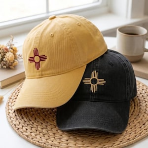 May include: A mustard yellow baseball cap with a maroon Zia symbol embroidery and a black baseball cap with a tan Zia symbol embroidery. Both caps feature a curved brim, a button on top, and stitched eyelets for ventilation on the crown.