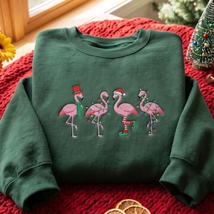 May include: A dark green crewneck sweatshirt featuring an embroidered graphic of four pink flamingos. The flamingos are decorated with festive elements including a red top hat, a green scarf, colorful string lights, and a Santa hat with striped elf socks.