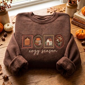 Puede incluir: Una sudadera marrón oscuro con marcos rectangulares y ovalados bordados que contienen una vela, una pila de libros con una calabaza, una casa y un ramo de flores. El texto "cozy season" está bordado en cursiva blanca debajo de los marcos.