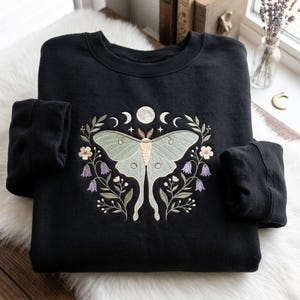 Op de afbeelding: Een zwart sweatshirt met ronde hals en een centrale borduursel van een lichtgroene maanspinner, omringd door paarse klokvormige bloemen, kleine witte bloesems, groene bladeren en een reeks maanfasen met een volle maan aan de bovenzijde.