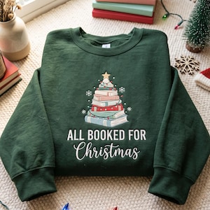 Peut inclure: Un sweat-shirt à col rond vert foncé avec un motif de sapin de Noël composé de livres empilés surmontés d'une étoile dorée, entouré de petits flocons de neige. Le texte "ALL BOOKED FOR Christmas" est imprimé en blanc sous le motif.