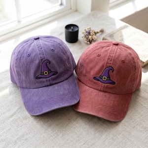 Può includere: Due cappellini da baseball, uno in viola slavato e uno in rosso slavato, ciascuno con un ricamo raffigurante un cappello da strega viola con una fascia verde e una fibbia gialla centrato sul pannello frontale della corona e visiera curva.