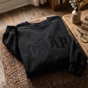 Op de afbeelding: Een zwart sweatshirt met lange mouwen en een ronde hals, voorzien van het woord GAAP geborduurd in bijpassende zwarte draad op de borst. Het kledingstuk heeft een geribbelde halslijn, boorden en zoom met een strak, effen ontwerp.
