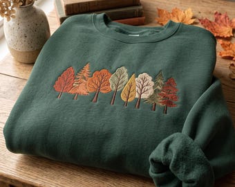 May include: A dark forest green crewneck sweatshirt featuring an embroidered row of various trees in autumn colours including orange, yellow, brown, and muted green. The embroidery is centred on the chest, showcasing detailed stitching of different leaf shapes.