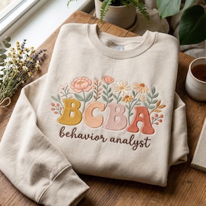 Pode incluir: Uma camisola de gola redonda cor de creme com texto bordado que diz BCBA behavior analyst numa mistura de tons amarelos, pêssego, rosa e laranja. O design é acentuado com caules florais, folhas e pequenas flores bordadas.