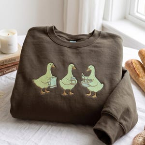 May include: A dark brown crewneck sweatshirt featuring an embroidered graphic of three light green geese. The first goose holds a towel, the second holds a jar, and the third holds a mixing bowl with a spoon. The design is centered on the chest.
