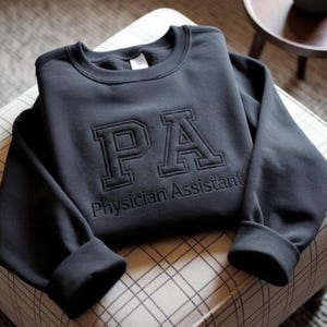 May include: A dark gray sweatshirt with the letters "PA" in a raised, block-letter style, and the words "Physician Assistant" below. The sweatshirt is folded on a patterned surface, with the sleeves cuffed.