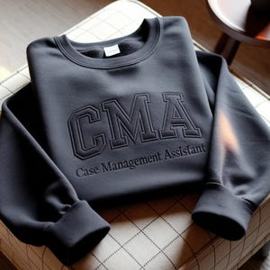 Könnte beinhalten: Ein dunkelgraues Sweatshirt mit den Buchstaben "CMA" in einem College-Stil gestickt, darunter "Case Management Assistant". Das Sweatshirt ist auf einem gemusterten Stuhl gefaltet.