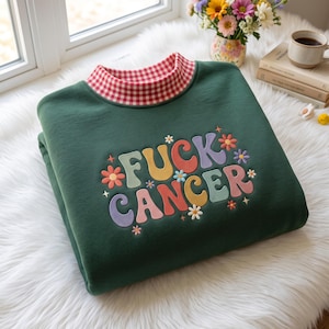 Op de afbeelding: Een donkergroen sweatshirt met een rood-witte vichy-ruitkraag. De voorkant toont de tekst FUCK CANCER in veelkleurige, afgeronde letters, omringd door kleine geborduurde bloemen in tinten oranje, roze, geel en blauw.