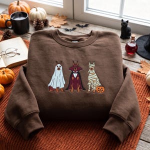 May include: A brown crewneck sweatshirt featuring an embroidered graphic of three Doberman dogs dressed in Halloween costumes. The first dog wears a white ghost sheet, the second a vampire cape with a bowtie, and the third is wrapped like a mummy.