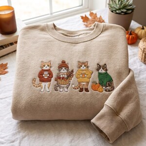 以下が含まれることがあります： 4匹の猫が刺繍されたベージュのクルーネックトレーナー。猫たちはセーターや小物を身につけており、茶色のセーター、落ち葉の入ったバスケットを持つ帽子をかぶった猫、マグカップを持った黄色のセーターの猫、緑色のセーターを着た猫が描かれています。