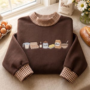 Op de afbeelding: Een donkerbruin sweatshirt met lange mouwen, voorzien van een bruin-witte gingham kraag en boorden. Het midden van het sweatshirt toont een geborduurde afbeelding van bakingrediënten, waaronder een maatbeker, eieren, vanille, suiker, boter en bloem.