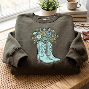 Könnte beinhalten: Ein olivgrünes Sweatshirt mit Rundhalsausschnitt und einer Stickerei von zwei hellblauen Cowboystiefeln. Die Stiefel sind mit einem bunten Strauß aus Wildblumen in Blau-, Gelb- und Weißtönen gefüllt, ergänzt durch grüne Stängel und Blätter.