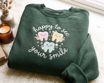 Puede incluir: Una sudadera de cuello redondo en color verde bosque oscuro con texto bordado en blanco que dice happy to see your smile. El diseño incluye tres dientes de dibujos animados en colores pastel rosa, amarillo y azul claro, rodeados de flores.