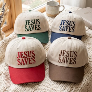 Op de afbeelding: Vier baseballpetten met een crèmekleurige kroon en contrasterende klep. Elke pet is voorzien van de geborduurde tekst 'JESUS SAVES' in een andere kleur: donkergroen, marineblauw, rood en bruin, passend bij de kleur van de klep.