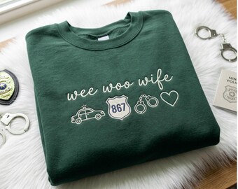 May include: A dark green crewneck sweatshirt featuring white embroidered text that reads "wee woo wife" above a line of icons including a police car, a badge with the number 867, a pair of handcuffs, and a heart shape, all in white thread.