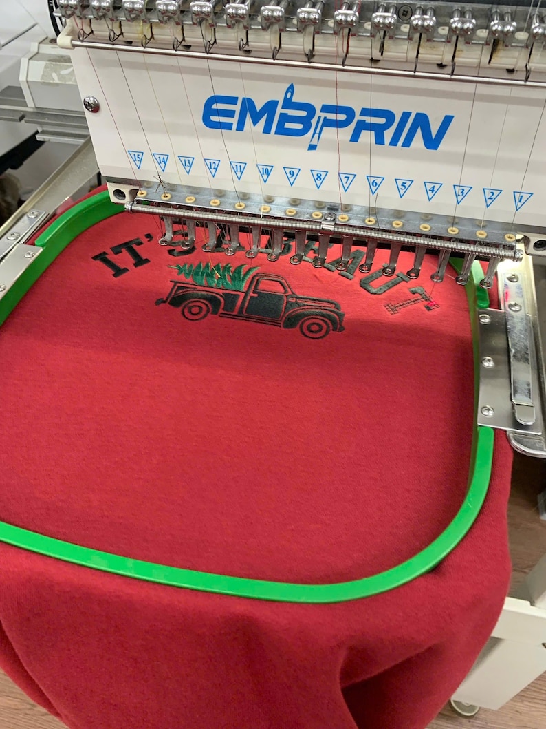May include: An Embprin embroidery machine with multiple needles stitching a design onto red fabric. The design features a black truck carrying a green pine tree with the text IT'S A BEAUT in black lettering, secured within a green embroidery hoop.