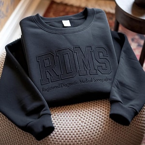 Peut inclure: Un sweat-shirt noir avec les lettres "RDMS" en police texturée de style universitaire. Le texte "Registered Diagnostic Medical Sonographer" est en dessous. Le sweat-shirt est plié sur une surface tissée.