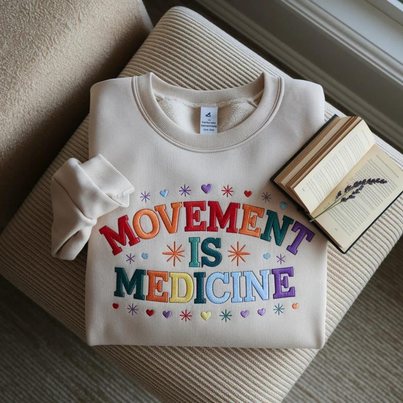 May include: Cream-colored sweatshirt with the embroidered text "MOVEMENT IS MEDICINE" in rainbow colors. The design includes hearts and stars. A book with lavender sprig rests nearby.