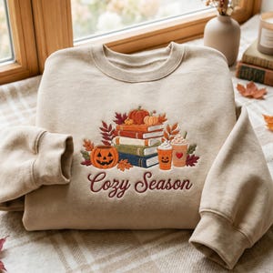 Pode incluir: Uma camisola bege de gola redonda com um design bordado de uma pilha de livros, abóboras e dois copos de café. O texto "Cozy Season" está bordado em letra cursiva vermelho-escura sob a pilha. O design inclui folhas de outono em tons terra.