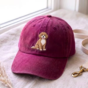 May include: A deep burgundy baseball cap featuring an embroidered graphic of a light brown dog with a white chest patch sitting upright. The dog has floppy ears, a black nose, and a small pink tongue visible. The cap has a curved brim and eyelets.