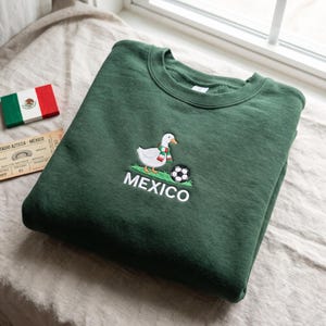 May include: A dark green crewneck sweatshirt featuring an embroidered graphic of a white goose wearing a green, white, and red striped scarf. The goose stands on a patch of green grass next to a black and white soccer ball with the word MEXICO below.