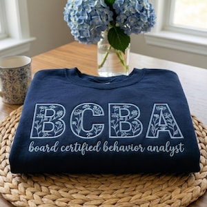 May include: A navy blue sweatshirt featuring the embroidered letters "BCBA" in white floral design. Underneath, the words "board certified behavior analyst" are written in a script font. The sweatshirt rests on a woven mat.