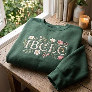 May include: A forest green crewneck sweatshirt featuring the embroidered white text IBCLC centered on the chest. The lettering is surrounded by detailed floral embroidery in shades of cream, soft pink, and sage green, including leaves and blossoms.