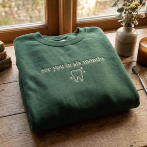 May include: A forest green crewneck sweatshirt featuring white embroidery on the chest. The text reads "see you in six months" above a line-art illustration of a tooth surrounded by four small four-pointed stars, all in white thread.