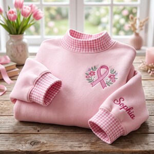 Op de afbeelding: Een lichtroze sweatshirt met een opstaande kraag en mouwboorden in een roze-wit vichy-ruitpatroon. Op de borst staat een geborduurd roze bewustzijnslintje omringd door kleine bloemen en blaadjes. De naam Sophia is op een van de mouwen geborduurd.