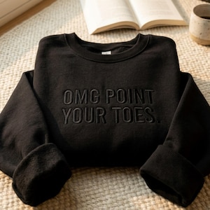 Puede incluir: Una sudadera negra de manga larga con el texto bordado "OMG POINT YOUR TOES." en hilo negro a juego. La prenda cuenta con cuello redondo, puños acanalados y un forro interior suave, presentada en un diseño limpio y sencillo.
