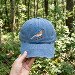Puede incluir: Una gorra de béisbol de color azul polvoriento con un diseño bordado de un pájaro azul con cabeza, alas y cola de color azul brillante, pecho naranja óxido y vientre blanco, posado sobre una pequeña rama marrón en el centro de la parte frontal.