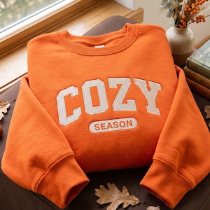 Peut inclure: Un sweat-shirt orange vif à col rond avec le mot COZY en grandes lettres majuscules crème sur la poitrine, et le mot SEASON brodé en orange à l'intérieur d'un cadre rectangulaire de couleur crème situé juste en dessous.