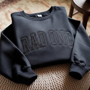 May include: A dark grey sweatshirt featuring the embroidered text "RAD ONC" in a collegiate style. The sweatshirt is neatly folded, highlighting the crew neck and ribbed cuffs. The garment is presented on a woven surface.