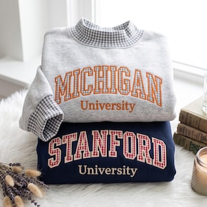 May include: A light grey sweatshirt with a mock neck and cuffs featuring a grey and white gingham pattern, displaying the orange embroidered text MICHIGAN University. Below it, a navy blue sweatshirt with red and white gingham lettering.
